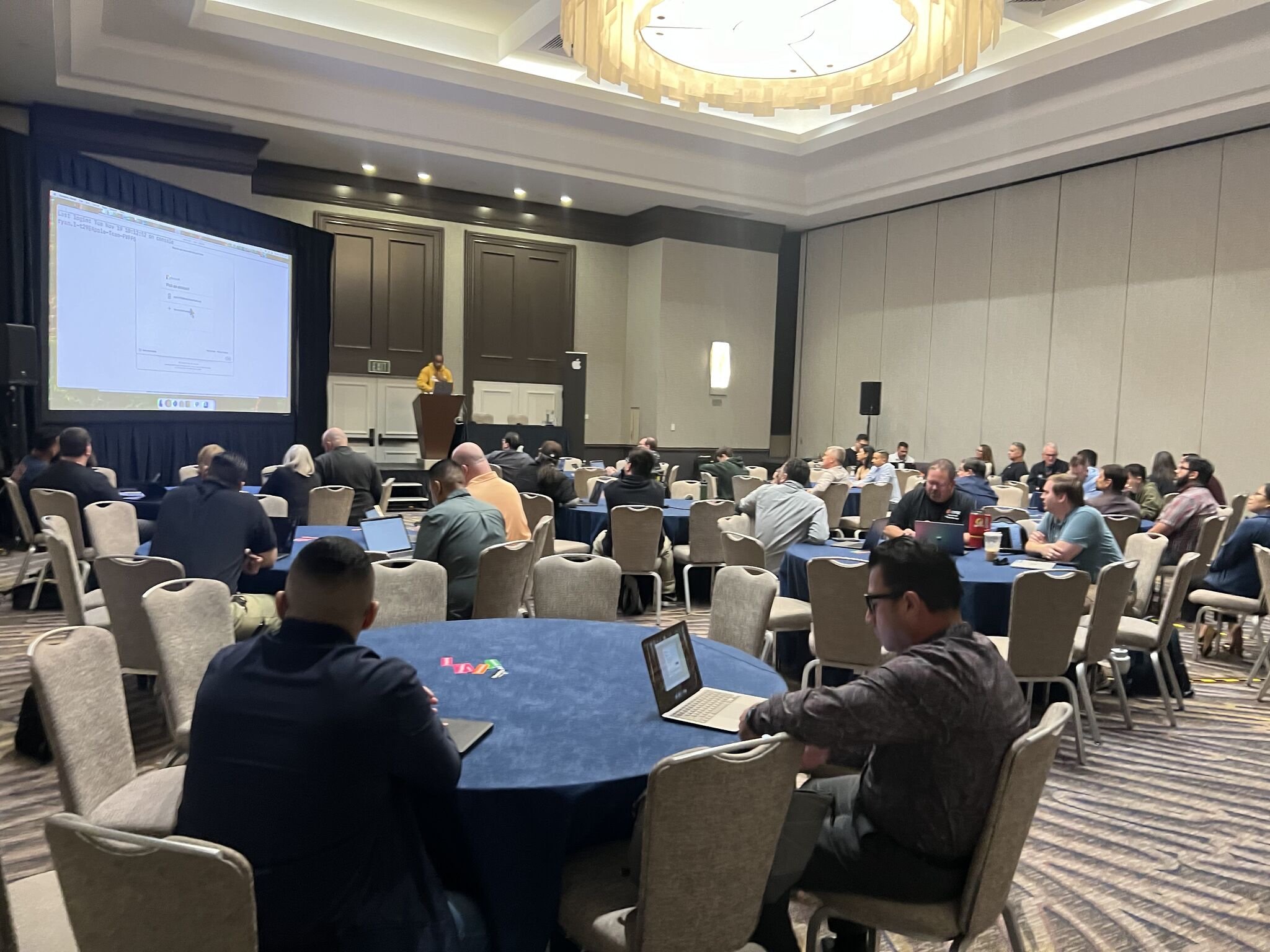 A full ballroom of round tables watching Kenan present from the podium at an Apple education event